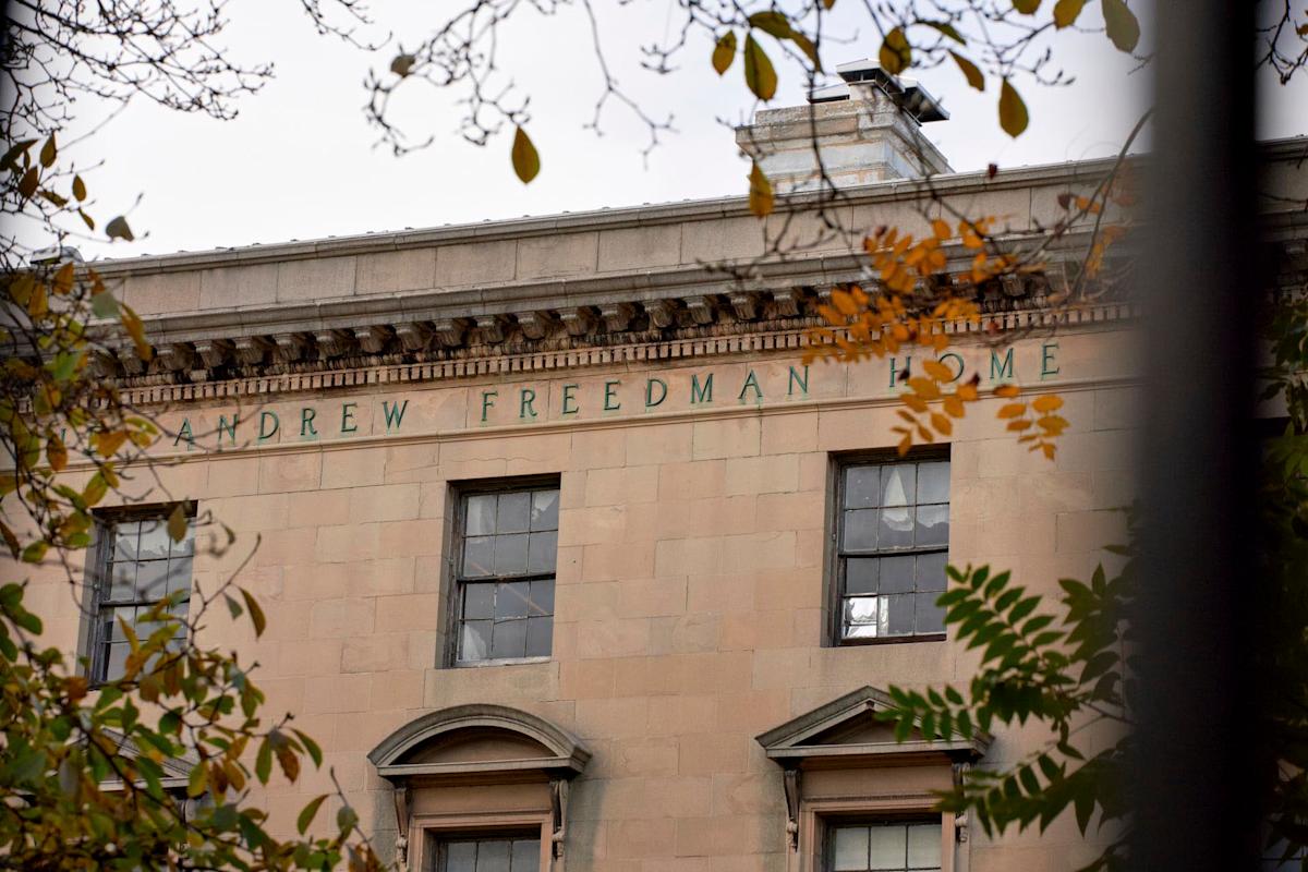 The upper facade of a stone building with green letters spelling "ANDREW FREEDMAN HOME," partially framed by autumn branches with yellow and green leaves.