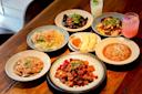 A wooden table set with various Chinese dishes, including spicy stir-fried chicken, lettuce salad, stir-fried eggplant, dumplings, noodles, and two colorful drinks, all served in ceramic bowls and plates.
