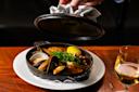 A hand lifts the lid off a cast iron skillet filled with seafood stew, including mussels, clams, and shrimp, garnished with greens and a lemon wedge, served on a white plate next to a glass of white wine.