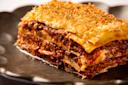 Close-up of a slice of lasagna on a dark plate. The layers of pasta, rich meat sauce, and melted cheese are topped with a golden breadcrumb crust, sprinkled with herbs, creating a deliciously appealing dish.