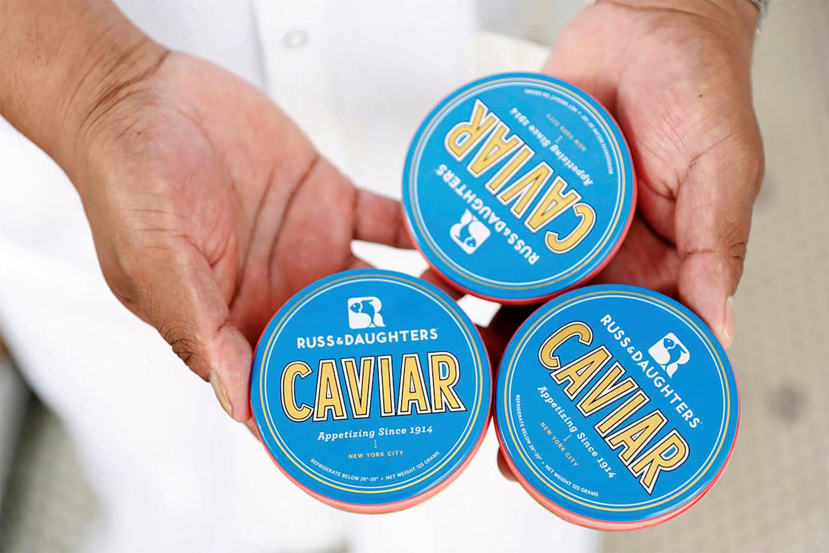 Hands holding three tins of Russ & Daughters caviar with blue lids and red rims. Each tin prominently displays "CAVIAR" and the company's logo.