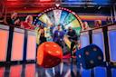 A colorful game show set with excited contestants standing at podiums, a spinning prize wheel in the background, and two large dice rolling toward the camera. The host stands in the center, smiling.