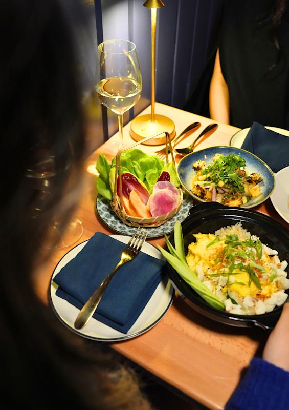 A table set with plates of assorted dishes, including a salad with lettuce and radishes, a bowl of scrambled eggs with scallions, and another dish with herbs. There is also a glass of white wine and navy napkins arranged on the plates.