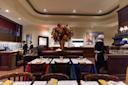 Elegant restaurant interior with tables covered in colorful tablecloths, a large floral arrangement in the center, staff in uniforms working, and a counter with glass display on the left. Warm lighting and artwork on the wall.
