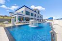 Large modern mansion with blue trim, featuring a luxurious swimming pool with a central fountain, surrounded by lounge chairs and white patio furniture under a clear blue sky.