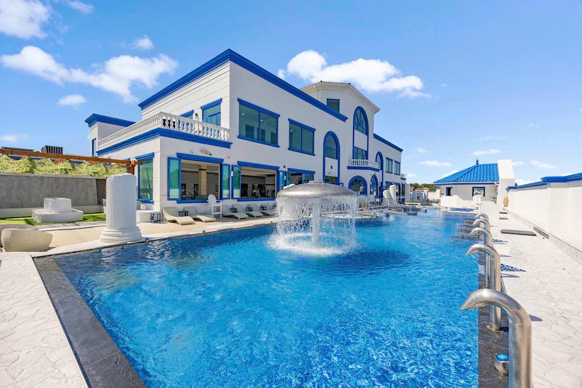 Large modern mansion with blue trim, featuring a luxurious swimming pool with a central fountain, surrounded by lounge chairs and white patio furniture under a clear blue sky.