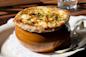 A bowl of French onion soup topped with melted, golden-brown cheese and sprinkled with chopped herbs, served on a white plate with a spoon and a glass of water in the background.