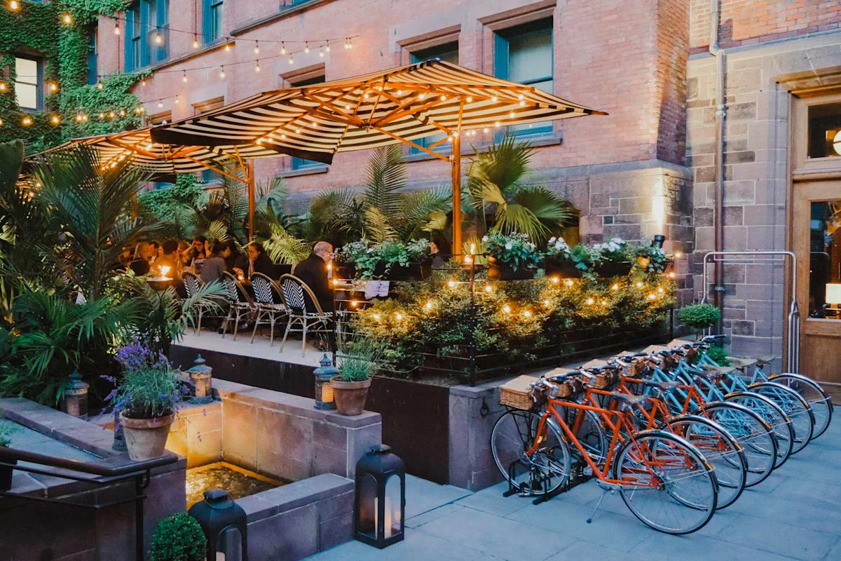 A cozy outdoor restaurant patio with string lights, striped awnings, lush plants, and patrons dining. Several orange and blue bicycles are parked nearby in front of a brick building.