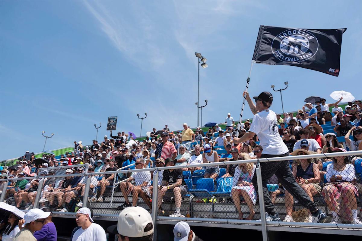 A man waves a  flag in front of a crowded outdoor stadium, where spectators sit in blue seats under a sunny sky, some using umbrellas and hats for shade.