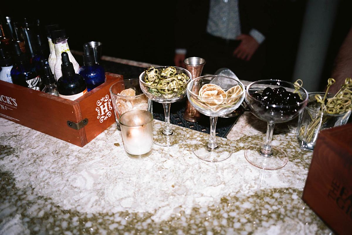 A bar counter with bottles, cocktail garnishes in coupe glasses, a candle, and a box of tools. A person in a suit is partially visible behind the counter.