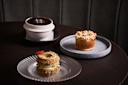 Three elegant desserts on a dark table: a layered pastry with cream on a glass plate, a pie topped with toasted meringue on a gray plate, and a round chocolate dessert in a white bowl with a tic-tac-toe design on top.