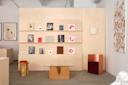 A modern room with a light wood display wall featuring books and art. A glass coffee table with a geometric cork base sits in front, with a wooden stool and a metallic chair beside it. Minimalist decor and neutral tones throughout.