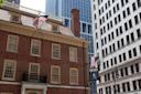 A historical brick building with American flags is surrounded by modern skyscrapers in a city. The building features colonial architecture with large windows and a prominent chimney.