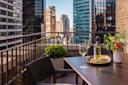 A modern balcony with a table set for two, featuring a bottle of white wine, two glasses, a vase of flowers, and a city skyline with tall buildings in the background.