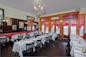 A cozy restaurant interior with wooden floors, red and white decor, and several tables covered in white tablecloths. Black chairs surround the tables, and a chandelier hangs from the ceiling. The walls feature mirrors and framed art.