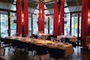 A dimly lit restaurant with large windows, red curtains, and chandeliers. Tables are elegantly set with white tablecloths, glassware, and tableware. Outside, trees and parked cars are visible through the windows.