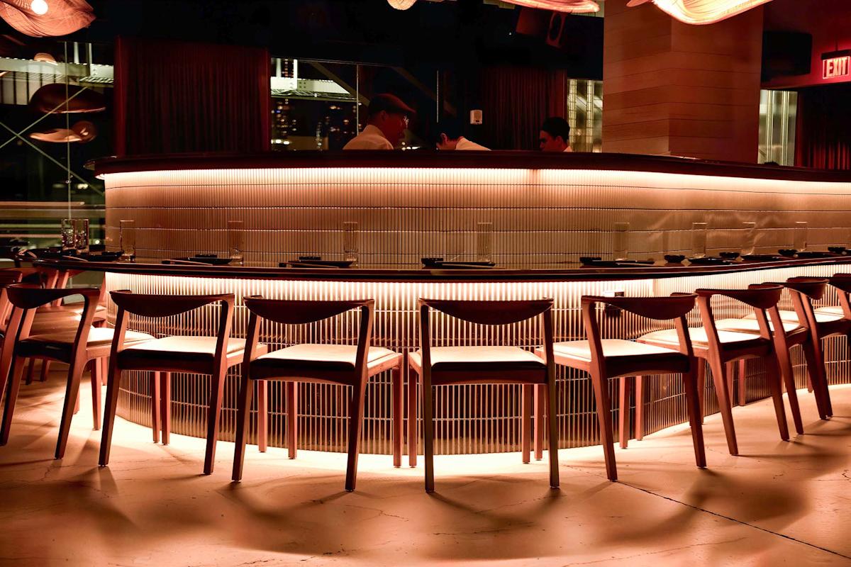 A modern, warmly lit bar with empty wooden chairs arranged in a semicircle, glowing under-counter lights, and two people standing behind the bar counter.