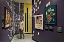 A man stands in a museum exhibit called "Streetscapes & Urban Oases," looking at framed posters and artwork on purple walls with city images and yellow geometric designs.