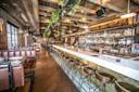 A rustic bar interior with a long white countertop, surrounded by metal stools. Wooden beams and plant decorations hang from the ceiling. Shelves stocked with bottles line the brick walls. Leather booths occupy the left side. Warm ambient lighting.