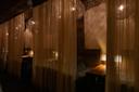 Dimly lit restaurant booths separated by sheer curtains, with wooden tables, cushioned benches, soft warm lighting, and rustic wooden and brick walls creating an intimate, cozy ambiance.