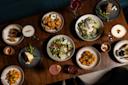 An overhead view of a wooden table set with assorted dishes, including salads, pasta, fried foods, bread, and various cocktails and desserts, arranged for a group meal.