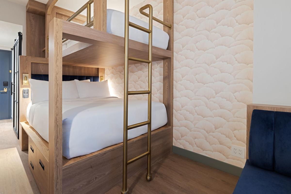 A modern hotel room with wooden bunk beds, white bedding, and a gold ladder. The walls have a soft, cloud-patterned wallpaper. A blue cushioned bench is partially visible on the right.