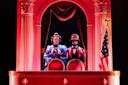 Two people in Victorian-era costumes stand in a theater box draped in red, under dramatic lighting, with columns, red curtains, and a U.S. flag to the right.