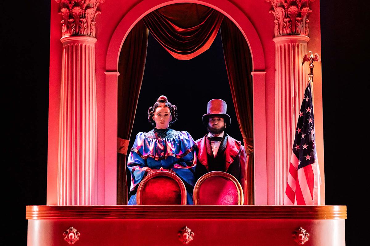 Two people in Victorian-era costumes stand in a theater box draped in red, under dramatic lighting, with columns, red curtains, and a U.S. flag to the right.