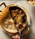 A hand holds a fork with a bite of biryani above a round dish filled with spiced rice and meat, topped with a baked bread crust, partially lifted to reveal the contents inside.