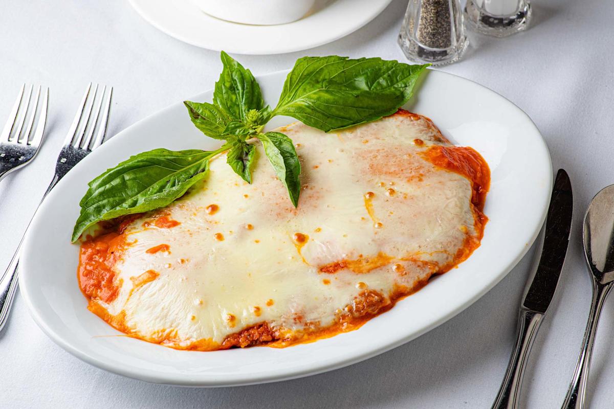 A plate of baked lasagna topped with melted cheese and garnished with fresh basil leaves, served on a white dish with cutlery and condiments on a white tablecloth.