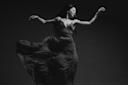 A black and white photo of a dancer gracefully moving. She is wearing a flowing dress that swirls around her, with arms elegantly extended. Her expression is focused and serene against a dark background.