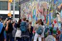 A group of people stand and observe a colorful, abstract mural on a street corner. They seem engaged, with some taking pictures. The mural features vibrant patterns and figures in motion. Nearby, a traffic light is visible.