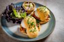 A plate with eggs Benedict topped with hollandaise sauce and chopped chives on English muffins, served with a leafy green salad and a small bowl of potato salad.