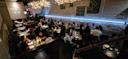 A modern, dimly-lit restaurant filled with people dining at tables. Overhead, decorative lights hang, and a mural is painted on a white brick wall. Guests are chatting, eating, and waitstaff are serving food.