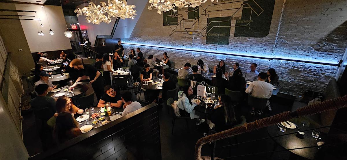 A modern, dimly-lit restaurant filled with people dining at tables. Overhead, decorative lights hang, and a mural is painted on a white brick wall. Guests are chatting, eating, and waitstaff are serving food.