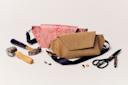 A crafting setup with a partially assembled brown handbag, red patterned fabric pieces, metal tools, a mallet, scissors, and small hardware parts on a white surface.