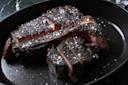 A sliced, charred steak with a bone, topped with coarse salt, is served in a black skillet. The inside of the steak is juicy and pink, contrasting with the dark, seared exterior.