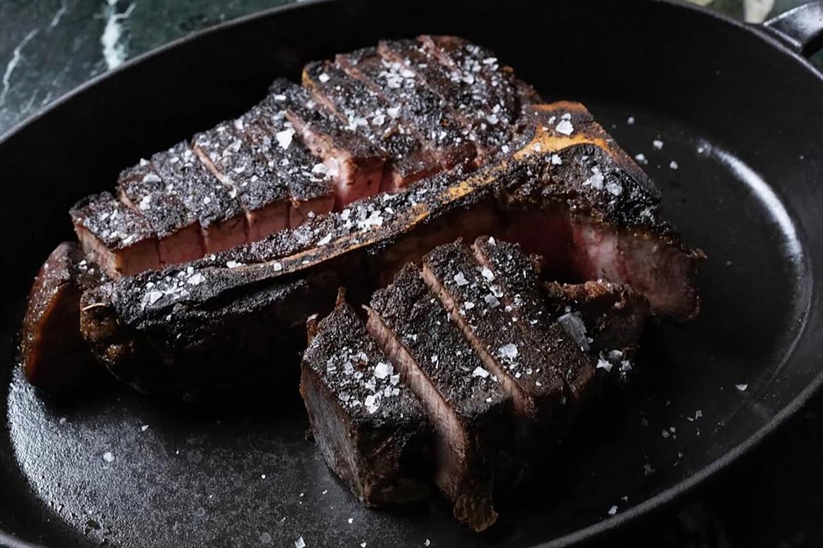 A sliced, charred steak with a bone, topped with coarse salt, is served in a black skillet. The inside of the steak is juicy and pink, contrasting with the dark, seared exterior.