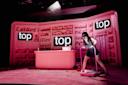 A woman in a black dress and headband stands on a treadmill in a pink room resembling a store, with shelves of products and two large screens displaying the word "top." A spotlight shines on her.