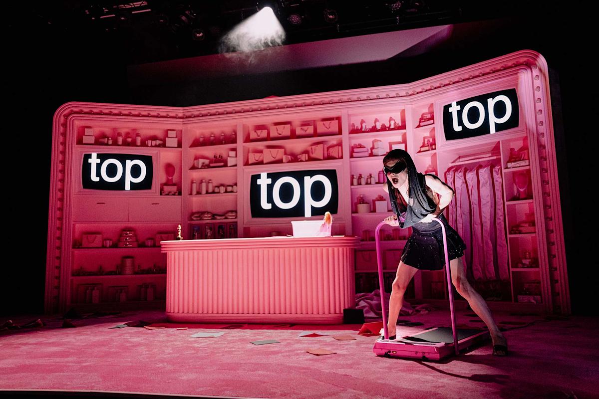 A woman in a black dress and headband stands on a treadmill in a pink room resembling a store, with shelves of products and two large screens displaying the word "top." A spotlight shines on her.
