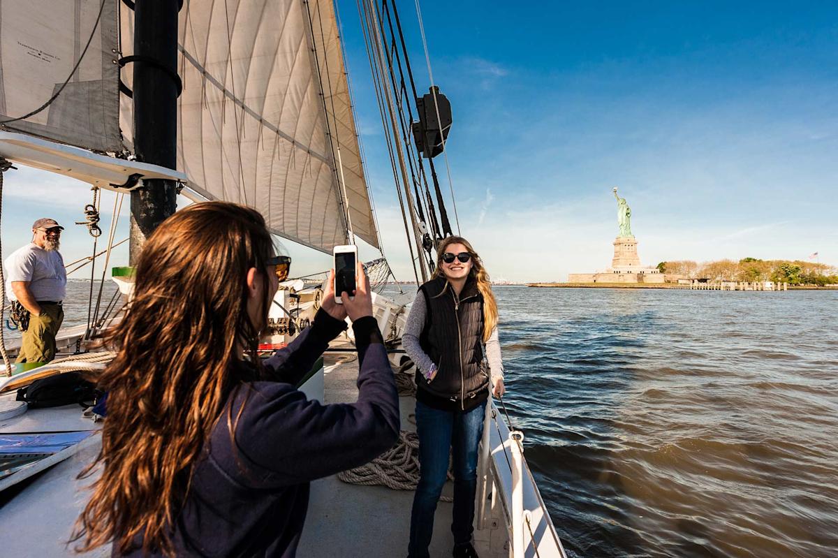 Two people pose for a photo on a sailboat with the Statue of Liberty in the background on a sunny day, while a person stands nearby on the boat.