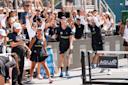 A group of pickleball players and supporters cheer enthusiastically at the side of an outdoor court during a match, with one woman in front smiling and raising her arms in celebration.