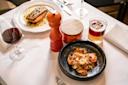 A table set with a plate of salmon, a black bowl of pasta with garnish, a glass of red wine, a cocktail, a red pepper grinder, and a wooden-lidded container on a white tablecloth.