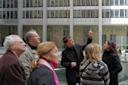 A group of people stand outdoors near a modern building with reflective glass. One person gestures upwards while the others look on attentively, suggesting they are observing or discussing something above.
