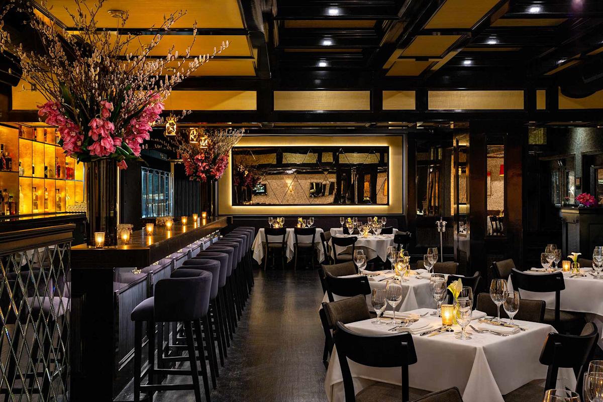 Elegant restaurant interior with dim lighting, tables set with white tablecloths and wine glasses, a long bar with high stools, floral arrangements, candles, and a large mirror on the wall creating a warm, inviting atmosphere.