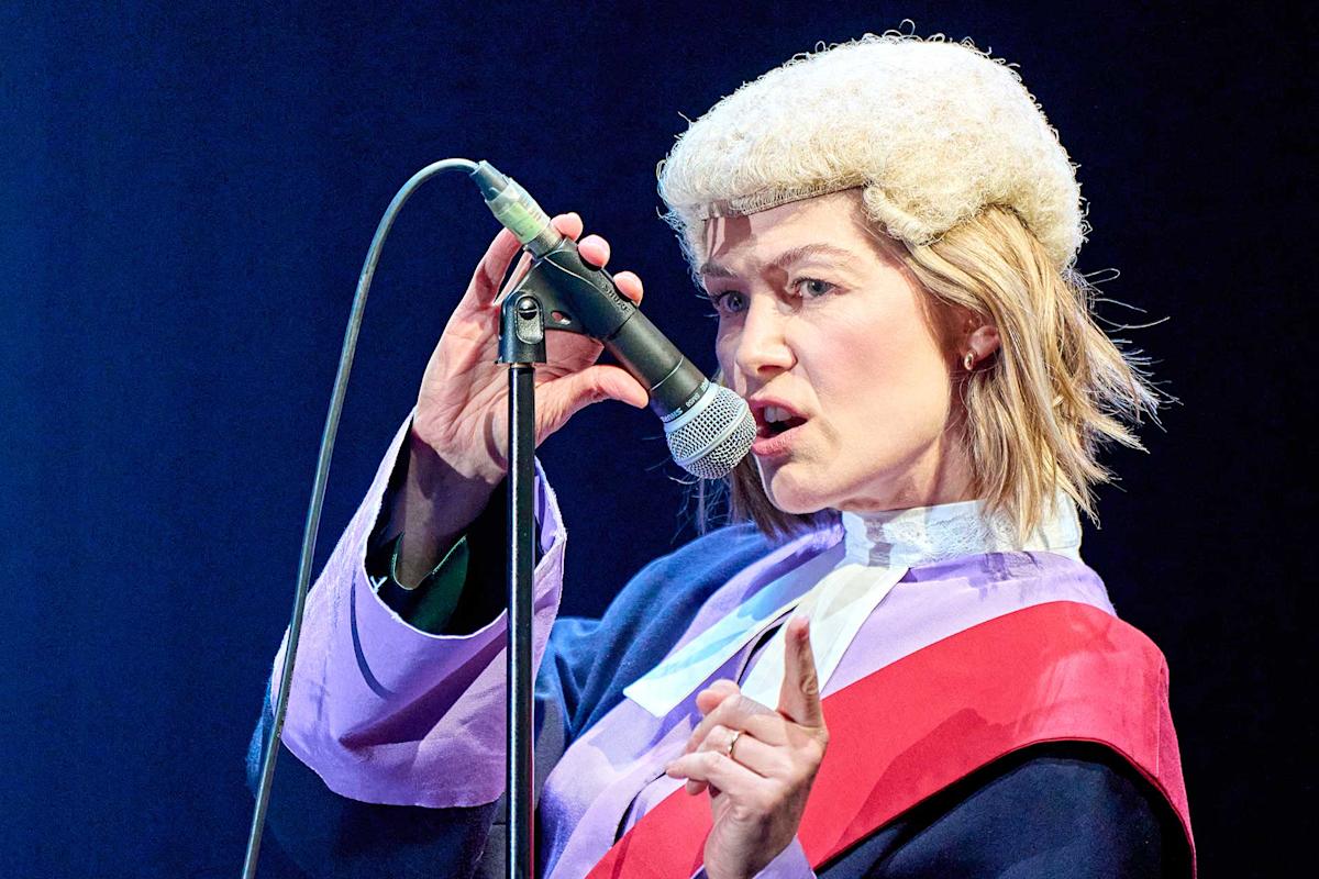 A person in a judge’s wig and robe speaks passionately into a microphone, holding up one finger as if making a point, against a dark background.