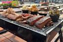 A tray of assorted barbecued meats, including ribs, brisket, and chicken, served with side dishes like coleslaw and pickles, is set on a picnic table at an outdoor restaurant.