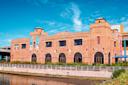 A large brick building with arched windows and castle-like turrets, situated by a waterfront under a bright blue sky. Banners hang on the left side of the building’s facade.