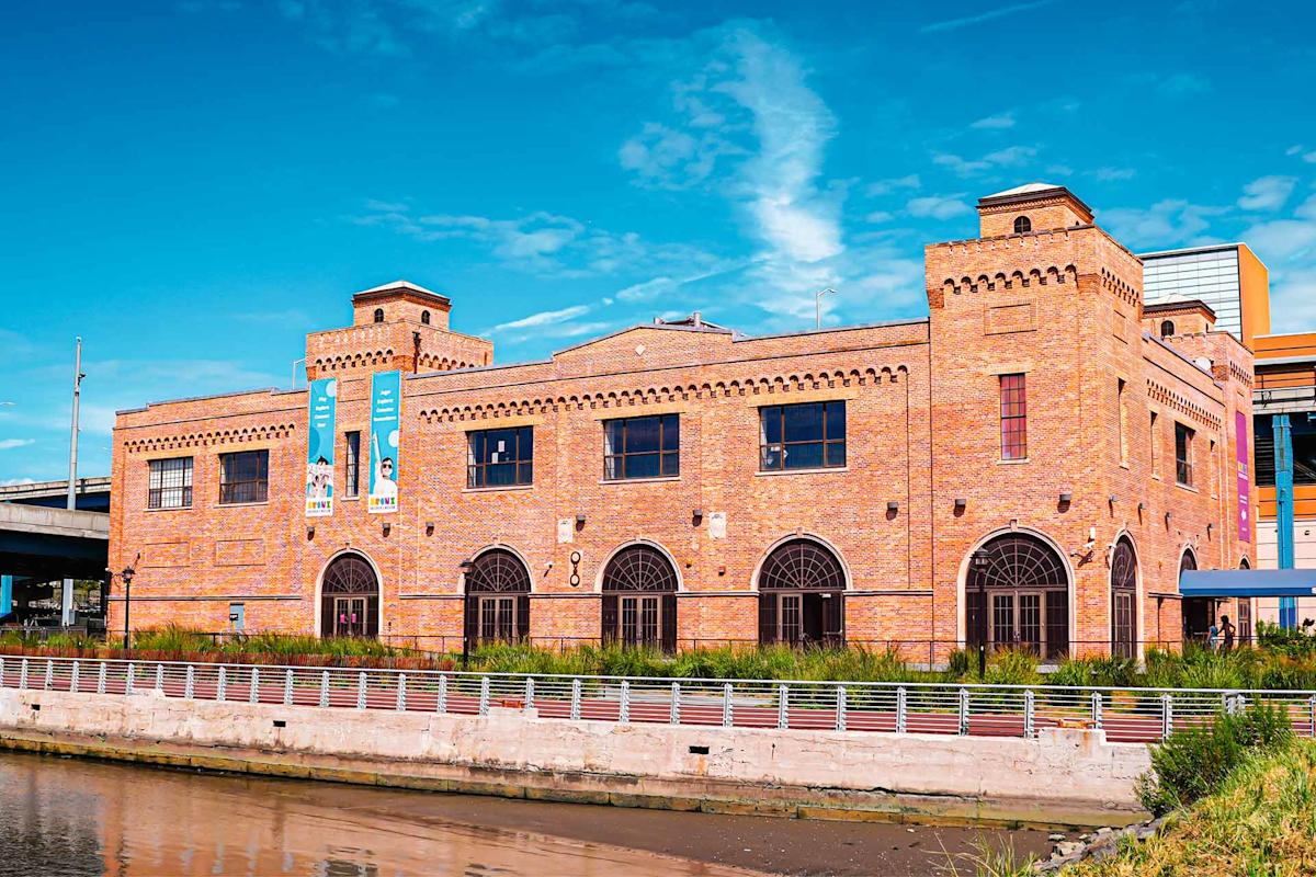 A large brick building with arched windows and castle-like turrets, situated by a waterfront under a bright blue sky. Banners hang on the left side of the building’s facade.
