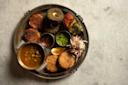 A traditional Indian thali on a metal plate featuring cutlets, kebabs, green chutney, dal in a bowl, grilled vegetables, sliced onions, a lemon wedge, and two small dipping sauces on a marble surface.